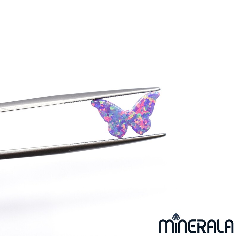Puede incluir: Un amuleto de mariposa de &oacute;palo morado y rosa con colores iridiscentes. El amuleto est&aacute; sujeto con unas pinzas.