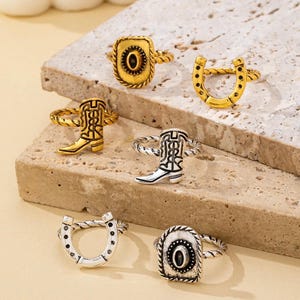 May include: A collection of six rings featuring western-inspired motifs. The set includes gold-toned rings with a horseshoe, a cowboy boot, and an oval emblem, alongside silver-toned rings with matching horseshoe, boot, and oval emblem designs.