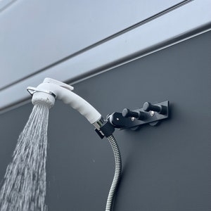 Może przedstawiać: Biała słuchawka prysznicowa z wypływającą wodą, zamontowana na czarnym wsporniku z trzema kołkami. Słuchawka prysznicowa jest połączona z elastycznym, srebrnym wężem. Tło to szara ściana.