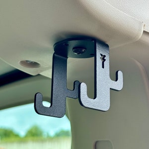 Pode incluir: Gancho de carro de metal preto com três ganchos pendurados no teto de um carro. O gancho é fixado ao teto com um parafuso.