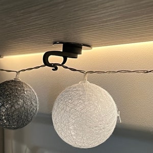 Puede incluir: Una luz de cadena blanca con un gancho negro unido a un techo blanco. La luz tiene dos luces de bola de algodón blancas y una luz de bola de algodón gris.