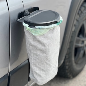 Può includere: Un bidone della spazzatura nero con un coperchio attaccato al lato di un veicolo argentato. Il bidone ha una fodera in tessuto grigio e un sacchetto per la spazzatura in plastica verde all'interno.