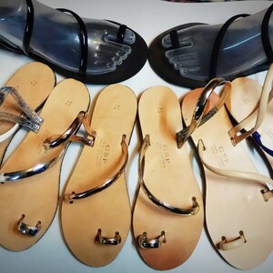 Puede incluir: Una colecci&oacute;n de sandalias con tiras de cuero y anillos para los dedos. Las sandalias son de varios colores, incluyendo blanco, tostado y azul. Las sandalias tienen un acento de metal dorado en las tiras.