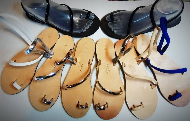 Puede incluir: Una colecci&oacute;n de sandalias con tiras de cuero y anillos para los dedos. Las sandalias son de varios colores, incluyendo blanco, tostado y azul. Las sandalias tienen un acento de metal dorado en las tiras.