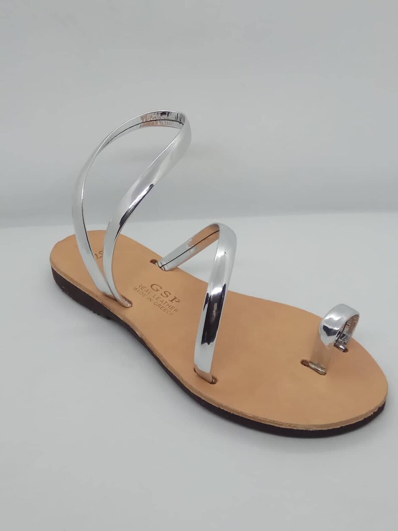 Puede incluir: Sandalias de cuero plateadas con un anillo para el dedo del pie y m&uacute;ltiples correas. Las sandalias est&aacute;n hechas en Grecia y tienen una suela de cuero marr&oacute;n.