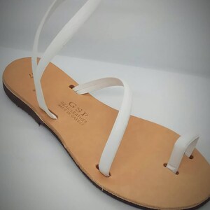 Puede incluir: Sandalias de cuero blanco con una sola correa sobre el pie y un bucle para el dedo del pie. Las sandalias est&aacute;n hechas en Grecia y tienen una suela de cuero marr&oacute;n.