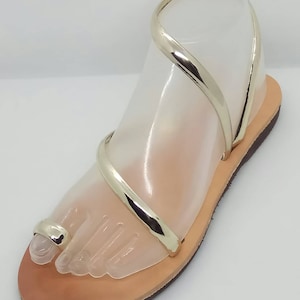 Puede incluir: Sandalias de cuero doradas con un anillo para el dedo del pie y tiras finas. Las sandalias tienen una suela plana y est&aacute;n hechas de cuero suave y flexible.