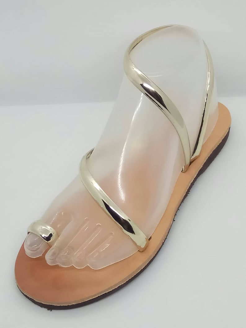 Puede incluir: Sandalias de cuero doradas con un anillo para el dedo del pie y tiras finas. Las sandalias tienen una suela plana y est&aacute;n hechas de cuero suave y flexible.