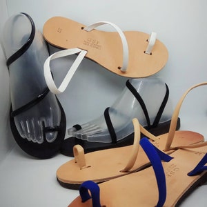 Puede incluir: Tres pares de sandalias de cuero con correas de diferentes colores. Las sandalias tienen un bucle para el dedo del pie y est&aacute;n hechas en Grecia. El texto en las sandalias dice "GSF Real Leather Made in Greece".