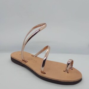 Puede incluir: Sandalias de cuero color oro rosa con un anillo para el dedo del pie y dos tiras que se envuelven alrededor del tobillo. Las sandalias tienen una suela de cuero marr&oacute;n.