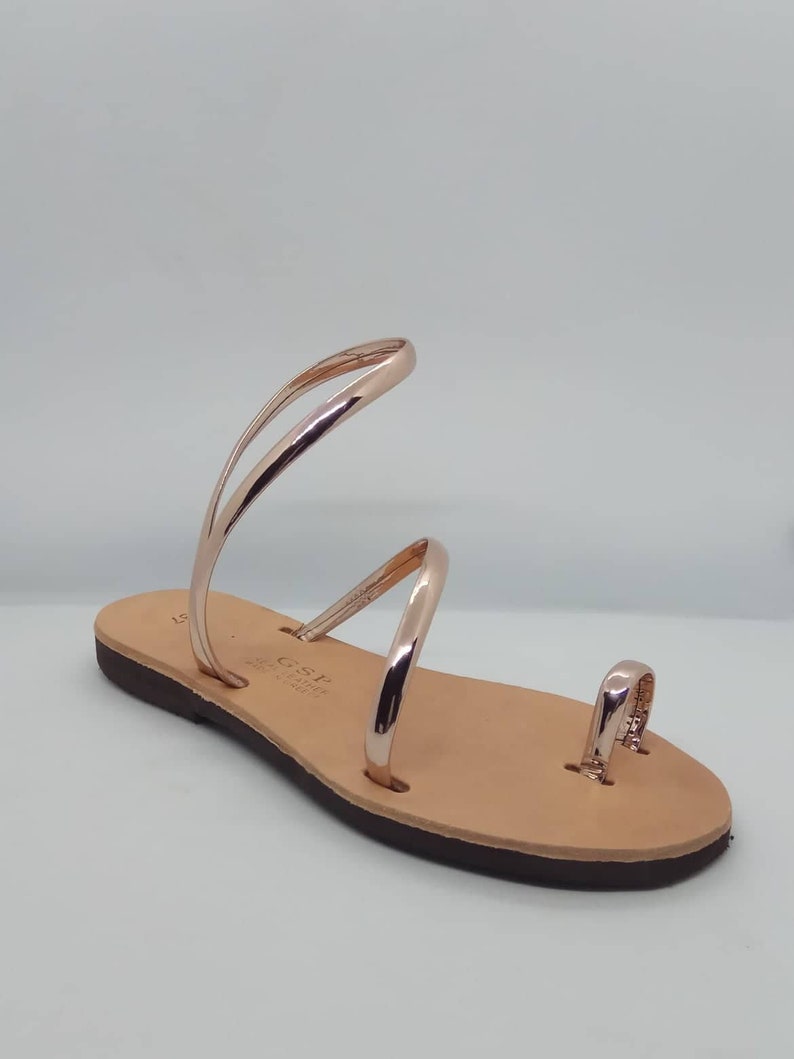 Puede incluir: Sandalias de cuero color oro rosa con un anillo para el dedo del pie y dos tiras que se envuelven alrededor del tobillo. Las sandalias tienen una suela de cuero marr&oacute;n.