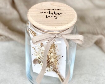 Könnte beinhalten: Ein Glasgefäß mit einem Holzdeckel, der mit einem beigefarbenen Band gebunden ist. Der Deckel trägt die Gravur "HAND IN HAND ein Leben lang". Das Gefäß enthält getrocknete Blumen.