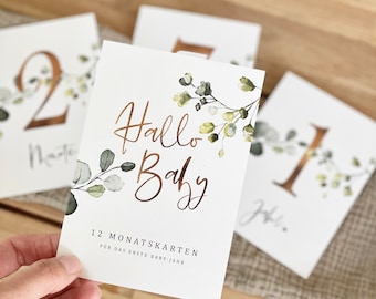 May include: Twelve cards with a watercolor floral design and gold foil lettering. The cards are numbered 1 through 12 and say "Hallo Baby" on the front. The text "12 Monatskarten für das erste Baby-Jahr" is printed on the bottom of the card.