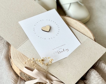 Puede incluir: Una tarjeta blanca con el texto "Eine kleine Überraschung für dich. Wir bekommen ein Baby." La tarjeta tiene un pequeño corazón de madera pegado a ella. La tarjeta está sobre una superficie de madera con un pequeño ramo de flores secas y un zapato de bebé blanco.