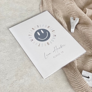 Könnte beinhalten: Eine weiße Karte mit einem blauen Smiley, der von einem Alphabetkreis umgeben ist. Der Text "Leon Hauber Klasse 1A" steht unter dem Smiley.