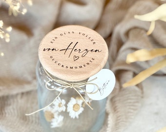 May include: A glass jar with a wooden lid that says "Ein Glas voller von Herzen Glücksmomente" with a small heart symbol. The jar is filled with white flowers and tied with twine.