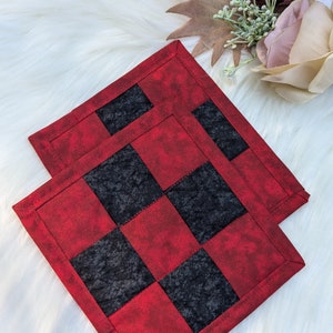 Op de afbeelding: Twee rode en zwarte geruite stoffen onderzetters. De onderzetters zijn gemaakt van een zachte, gestructureerde stof en hebben een subtiele vintage uitstraling.