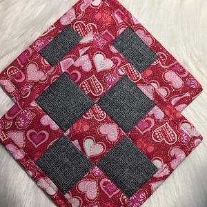 Peut inclure: Deux dessous de plat carrés matelassés. Les dessous de plat présentent un fond rouge avec un motif de cœurs roses et blancs. Chaque dessous de plat comporte quatre carrés gris foncé. Les bords sont bordés d'une bordure rouge.