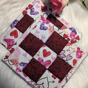 May include: Two red and white quilted pot holders with a heart pattern. The pot holders are made of fabric with a patchwork design.