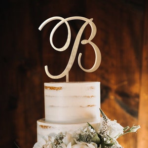 May include: A two-tiered white cake with a gold-colored letter "B" cake topper. The cake is decorated with white flowers and greenery. The cake has a rustic, semi-naked design.