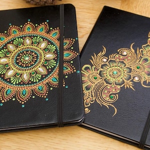 Pode incluir: Dois diários de couro preto com desenhos pintados à mão. Um diário apresenta um mandala colorido com detalhes verdes, amarelos e laranja. O outro diário apresenta um design floral com detalhes dourados e vermelhos.