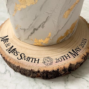 May include: A wooden cake stand with a natural bark edge, engraved with "Mr. & Mrs. Smith" and "20th May 2023". A 20-sided die is also engraved on the stand.