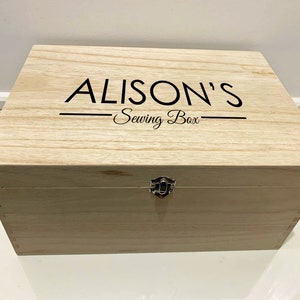 Peut inclure: Une bo&icirc;te en bois avec un couvercle &agrave; charni&egrave;res et un fermoir en m&eacute;tal. La bo&icirc;te est de couleur brun clair et porte l'inscription "ALISON'S Sewing Box" sur le dessus.