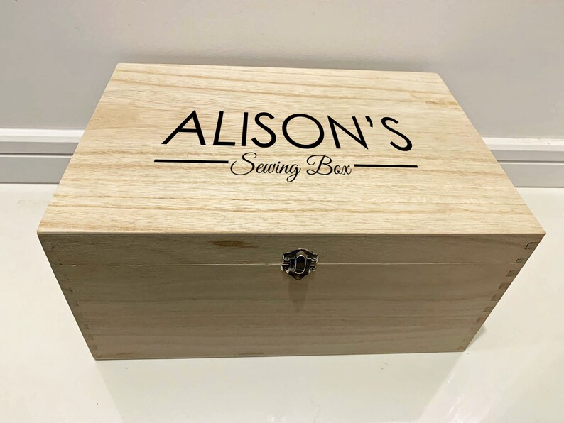 Peut inclure: Une bo&icirc;te en bois avec un couvercle &agrave; charni&egrave;res et un fermoir en m&eacute;tal. La bo&icirc;te est de couleur brun clair et porte l'inscription "ALISON'S Sewing Box" sur le dessus.