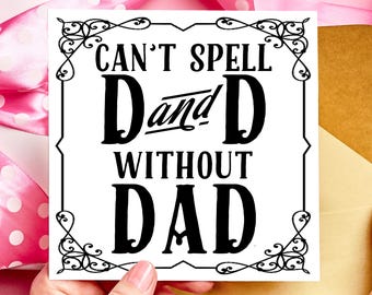 May include: A white greeting card with black text that reads "Can't spell Dad without Dad" in a decorative frame. The card is held against a pink and gold background with pink ribbon.