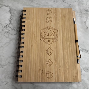 Puede incluir: Un cuaderno de espiral de madera con un portaplumas y un diseño de un dado de 20 caras con los números del 1 al 20 en las caras.