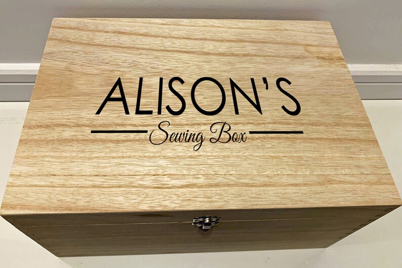 Peut inclure: Une bo&icirc;te en bois avec un couvercle &agrave; charni&egrave;res et un fermoir en m&eacute;tal. La bo&icirc;te est &eacute;tiquet&eacute;e "ALISON'S Sewing Box" en lettres noires.