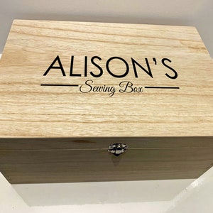 Peut inclure: Une bo&icirc;te en bois avec un couvercle &agrave; charni&egrave;res et un loquet en m&eacute;tal. La bo&icirc;te est de couleur brun clair et porte l'inscription "ALISON'S Sewing Box" sur le dessus.