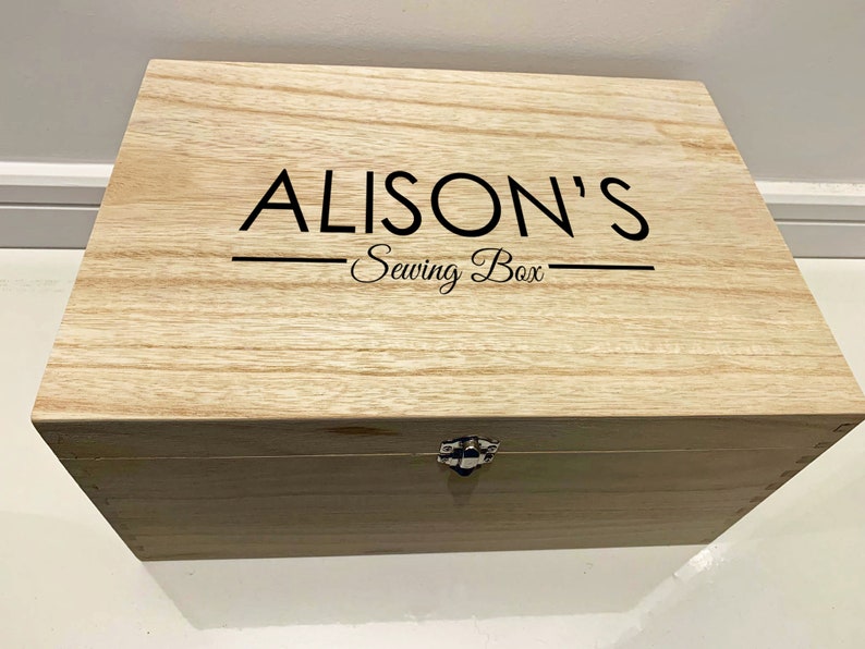 Peut inclure: Une bo&icirc;te en bois avec un couvercle &agrave; charni&egrave;res et un loquet en m&eacute;tal. La bo&icirc;te est de couleur brun clair et porte l'inscription "ALISON'S Sewing Box" sur le dessus.