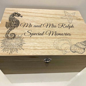 Może przedstawiać: Drewniana skrzynia z pokrywą na zawiasach, ozdobiona konikiem morskim, gwiazdą morską i muszelkami. Skrzynia jest grawerowana tekstem "Mr and Mrs Ralph Special Memories".