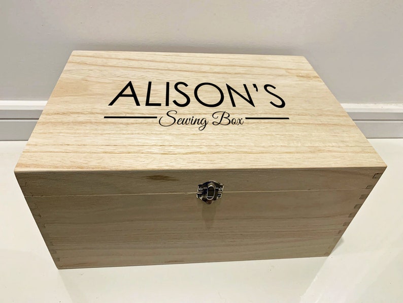 Peut inclure: Une bo&icirc;te en bois avec un couvercle &agrave; charni&egrave;res et un loquet en m&eacute;tal. La bo&icirc;te est de couleur brun clair et porte la mention "ALISON'S Sewing Box" grav&eacute;e sur le dessus.