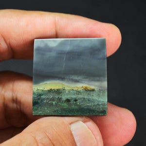 Op de afbeelding: Een vierkante, gepolijste steencabochon met een landschapsachtig patroon, voorzien van een donkergrijs bovengedeelte, een dunne horizontale band in mosterdgeel en een textuurrijke groen en donkergroene basis die doet denken aan een horizonlijn.