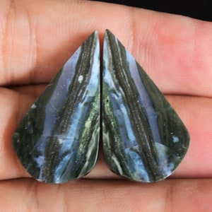 May include: Two polished, teardrop-shaped gemstones with a mix of dark green, black, and blue hues. The stones have a striped pattern and are held in a hand against a black background. These stones could be used for jewelry.