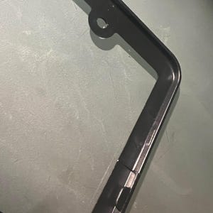 May include: Black metal license plate frame with a hole at the top for hanging. The frame has a smooth, glossy finish.