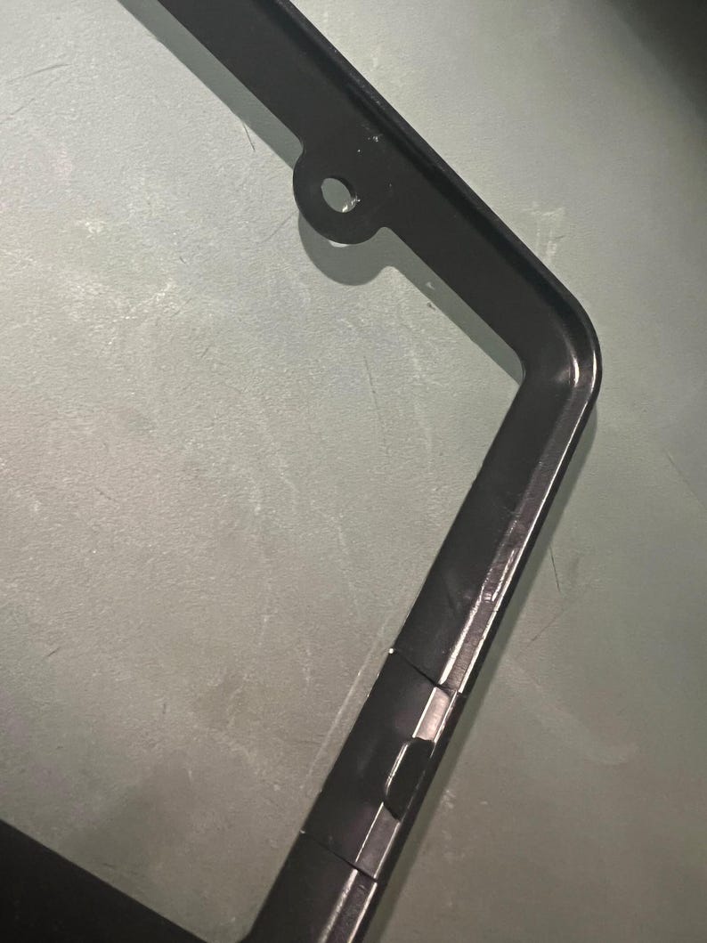 May include: Black metal license plate frame with a hole at the top for hanging. The frame has a smooth, glossy finish.