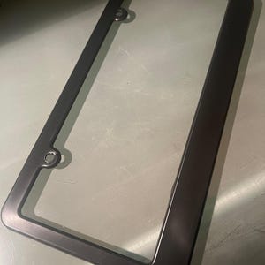 May include: Black plastic license plate frame with rounded corners and two holes on the top and bottom.