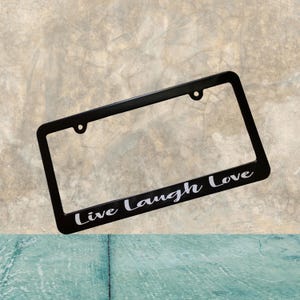 Può includere: Cornice nera per targa con le parole "Live Laugh Love" in corsivo bianco. La cornice ha una forma rettangolare con angoli arrotondati e due fori di montaggio nella parte superiore.