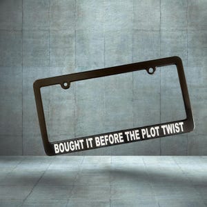 May include: Black license plate frame with the text "BOUGHT IT BEFORE THE PLOT TWIST" in white. The frame has a rectangular shape with rounded corners and two mounting holes at the top.