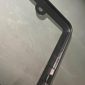 May include: Black metal license plate frame with a hole at the top for hanging. The frame has a smooth, glossy finish.