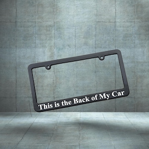 Puede incluir: Marco de matrícula negro con el texto "This is the Back of My Car" en blanco. El marco tiene dos orificios de montaje y una forma rectangular. El fondo es una pared de hormigón gris.