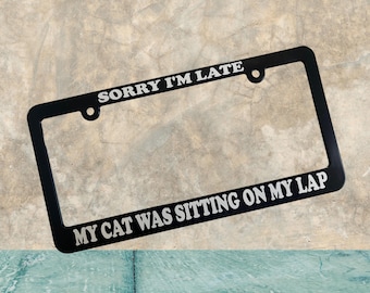 May include: Black metal license plate frame with white text that reads "Sorry I'm Late My Cat Was Sitting On My Lap".