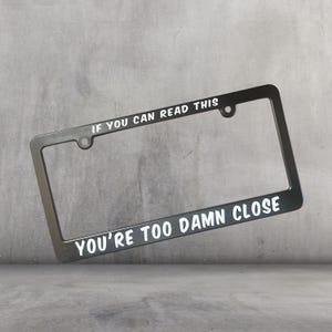 Puede incluir: Marco de matrícula negro con texto blanco: "IF YOU CAN READ THIS YOU'RE TOO DAMN CLOSE". El marco es rectangular, con esquinas redondeadas y orificios de montaje.