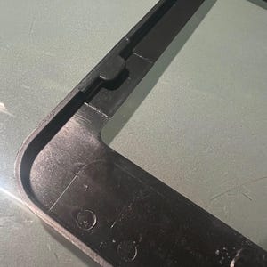 May include: Black plastic frame with rounded corners and two small holes. The frame is designed to hold a license plate.
