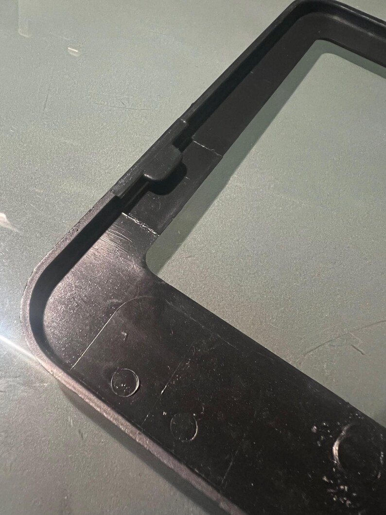 May include: Black plastic frame with rounded corners and two small holes. The frame is designed to hold a license plate.