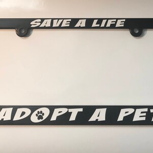 May include: Black plastic license plate frame with white text that reads "Save a Life Adopt a Pet" with a paw print icon.