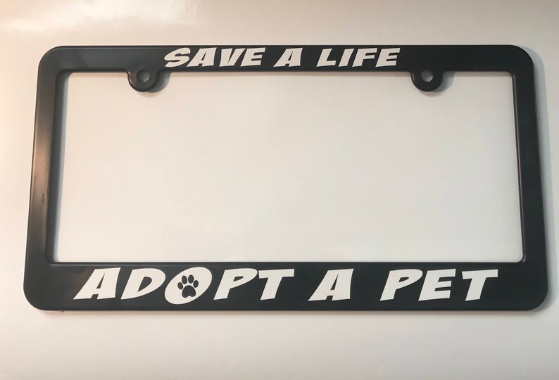 May include: Black plastic license plate frame with white text that reads "Save a Life Adopt a Pet" with a paw print icon.